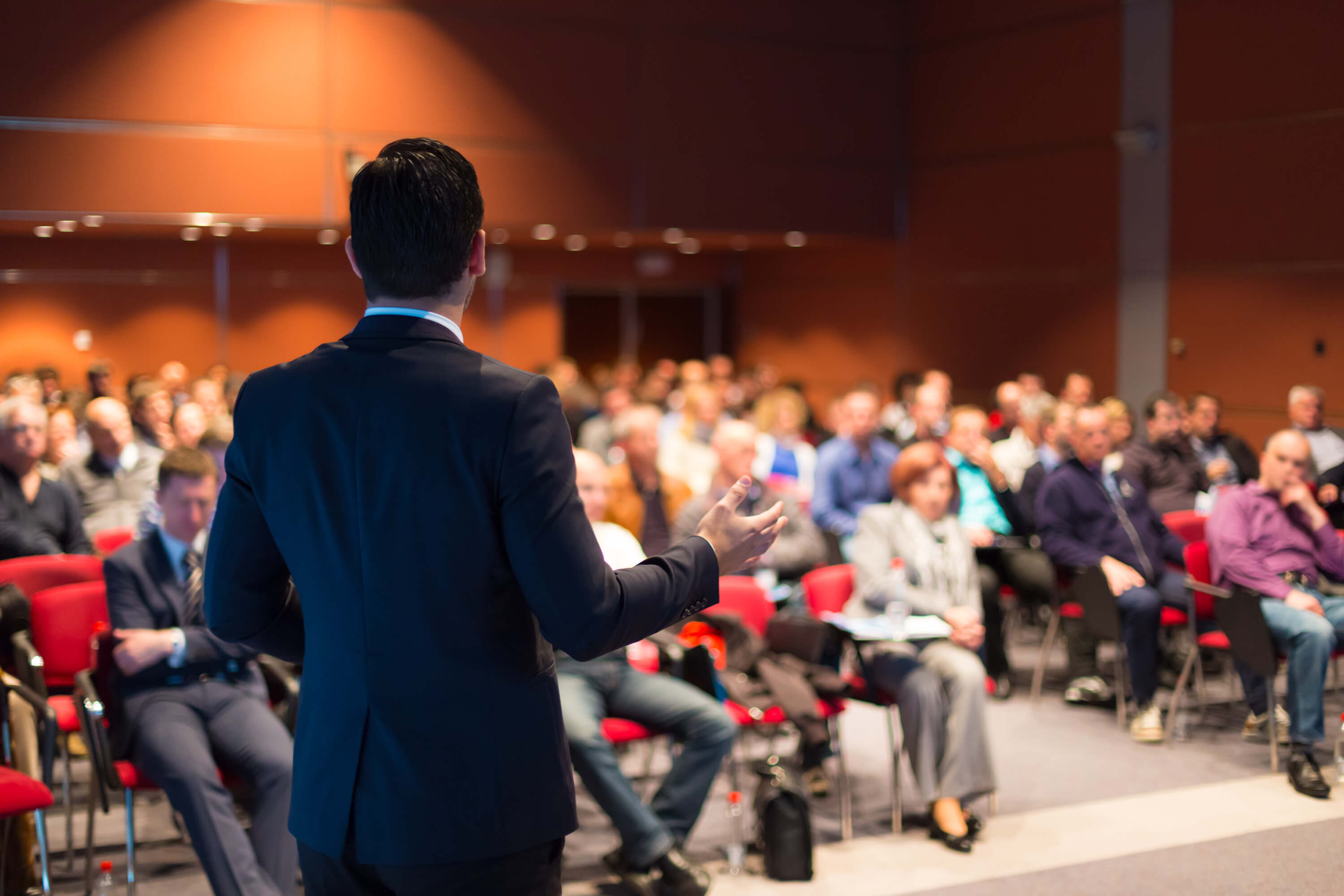 In the blurred background people are in a conference and a speaker delivers a speech.